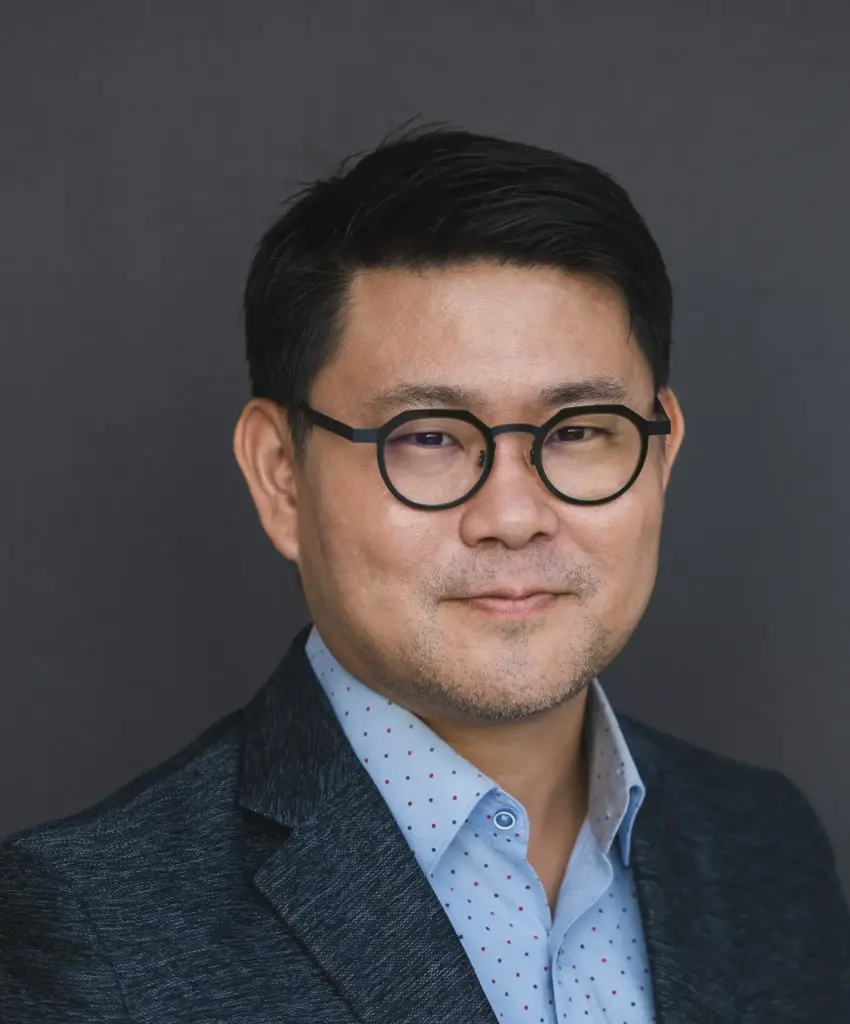 Professional head-and-shoulders portrait of a man with round black glasses wearing a blue polka-dotted shirt and dark blazer, neutral expression, dark background.